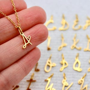 May include: A gold-colored necklace with a pendant shaped like the letter "A" and a small heart. The pendant is attached to a delicate gold chain. Other gold letter pendants are visible in the background.