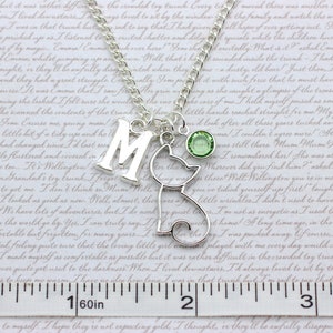 May include: Silver charm necklace with a letter M, a cat charm, and a green crystal. The necklace is on a white background with a ruler.