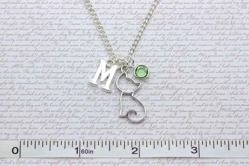 May include: Silver charm necklace with a letter M, a cat charm, and a green crystal. The necklace is on a white background with a ruler.