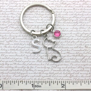 May include: Silver keychain with a cat charm, a letter 'S' charm, and a pink crystal charm. The keychain is attached to a silver key ring. The background is a piece of paper with text.