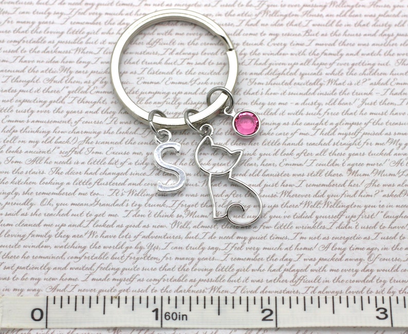 May include: Silver keychain with a cat charm, a letter 'S' charm, and a pink crystal charm. The keychain is attached to a silver key ring. The background is a piece of paper with text.