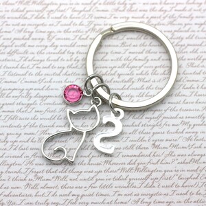 May include: A silver keychain with a pink rhinestone charm, a silver cat charm, and a silver number 2 charm. The keychain is on a white background with text.