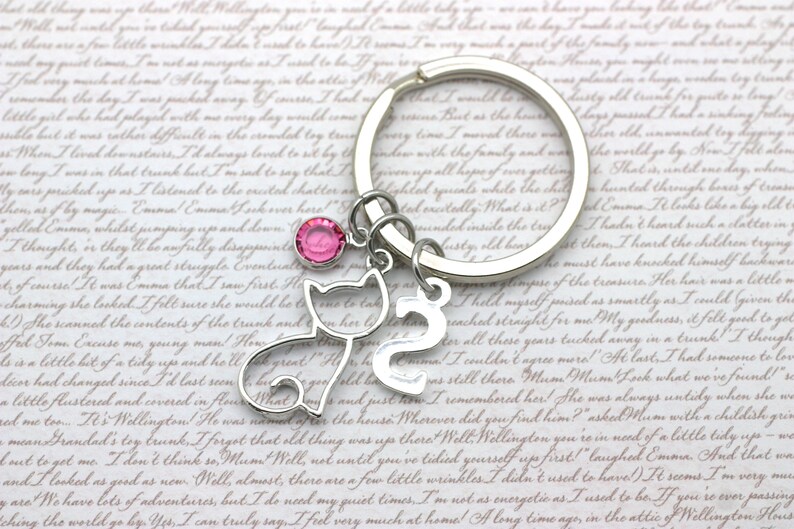 May include: A silver keychain with a pink rhinestone charm, a silver cat charm, and a silver number 2 charm. The keychain is on a white background with text.