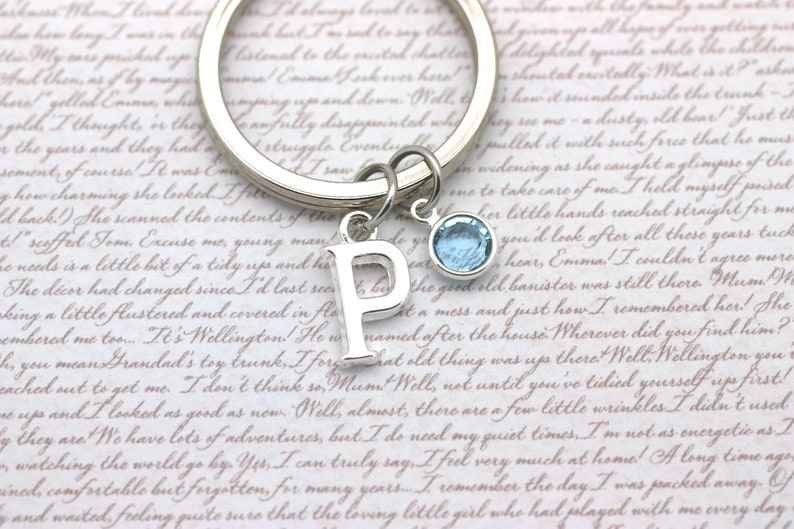 May include: Silver keyring with a charm of the letter P and a light blue crystal charm. The keyring is on a piece of paper with text.