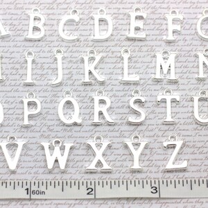 May include: Silver alphabet letter charms, A through Z, with a hole at the top for hanging. The charms are arranged in alphabetical order on a white surface with a ruler.