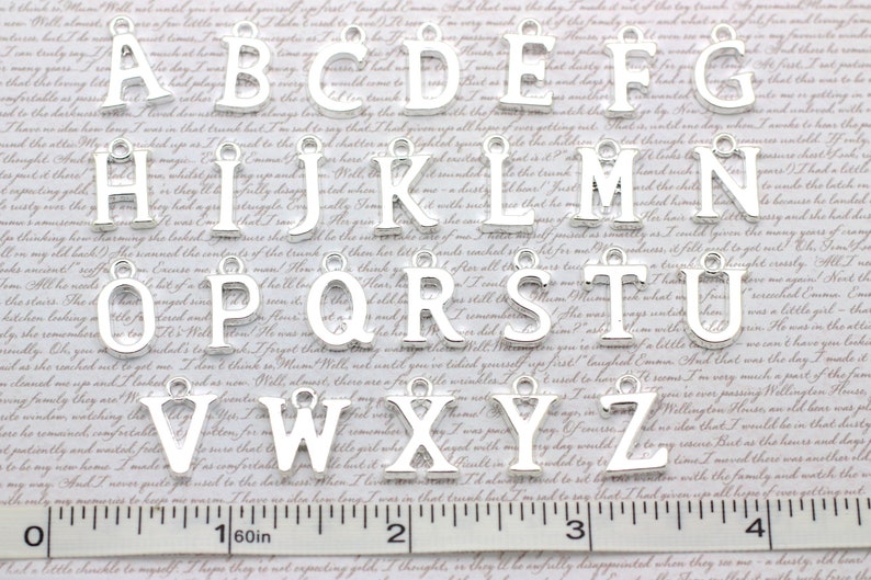 May include: Silver alphabet letter charms, A through Z, with a hole at the top for hanging. The charms are arranged in alphabetical order on a white surface with a ruler.