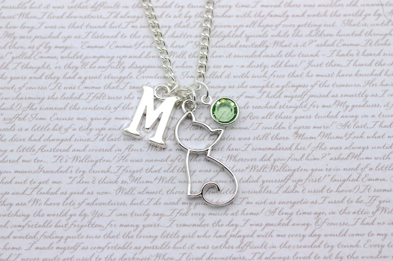 May include: A silver charm necklace with a letter 'M' charm, a silver cat charm, and a light green crystal charm. The necklace is on a white background with text.