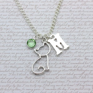 May include: A silver necklace with a charm of a cat, a charm of the letter 'M', and a green crystal charm. The necklace is on a white background with text.