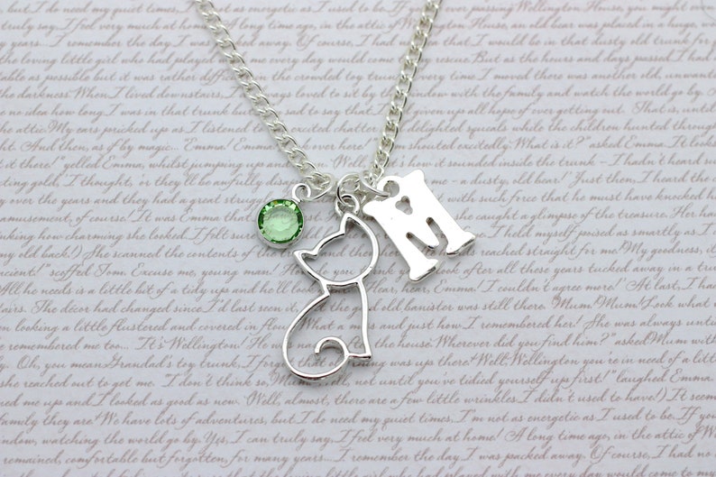 May include: A silver necklace with a charm of a cat, a charm of the letter 'M', and a green crystal charm. The necklace is on a white background with text.