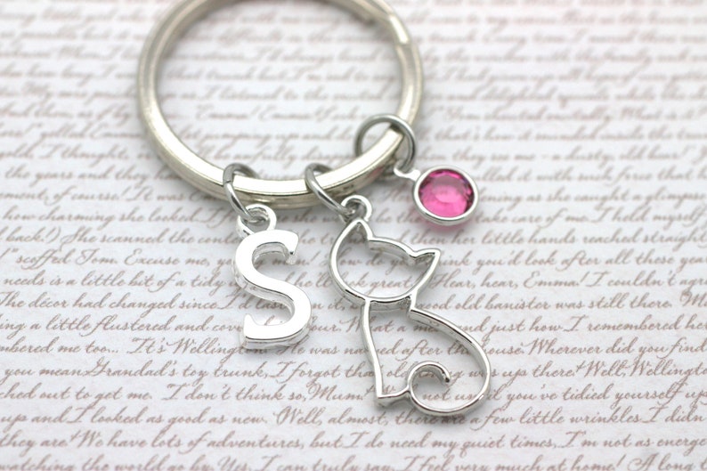May include: A silver keychain with a letter 'S' charm, a small cat charm, and a pink rhinestone charm. The keychain is on a white background with cursive writing.