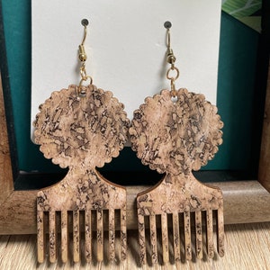 May include: A pair of wooden earrings with a brown and black marbled pattern. The earrings are shaped like an afro with a comb underneath. The earrings are labeled "Echo Earth Jewelry".