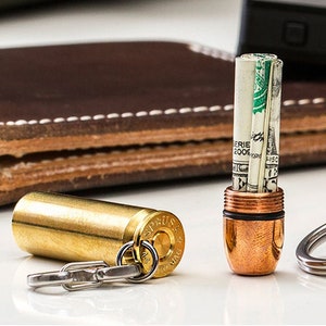 May include: A brass bullet-shaped money holder with a keychain attached. The holder is open, revealing rolled-up banknotes. A second holder, also bullet-shaped, is closed. A brown leather wallet is in the background.