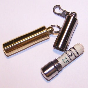 May include: A gold-colored metal keychain money holder with a silver-colored metal cap and a silver-colored metal carabiner clip. A rolled-up five-dollar bill is visible inside the open holder. The holder is designed to keep money safe and secure.