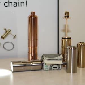 May include: A collection of metallic keychains and accessories. The image features a copper-colored cylindrical keychain, a silver flashlight, and other gold and silver metal components. A roll of US currency is placed near the flashlight.