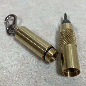 May include: A disassembled brass container with a textured grip and a silver carabiner clip. The container is cylindrical with a threaded opening and a black O-ring seal. The other end holds a dark-colored tool.