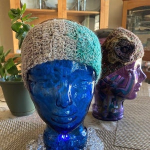 May include: A hand-crocheted hat in grey and teal sits on a blue glass head. Another hat is displayed on a purple glass head. Clear, faceted crystals surround the base of the blue head. The hats are handmade.