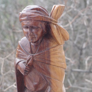 Puede incluir: Una escultura de madera de una persona que lleva una cubierta para la cabeza y una capa. La escultura est&aacute; tallada en un estilo realista y tiene un color marr&oacute;n c&aacute;lido.