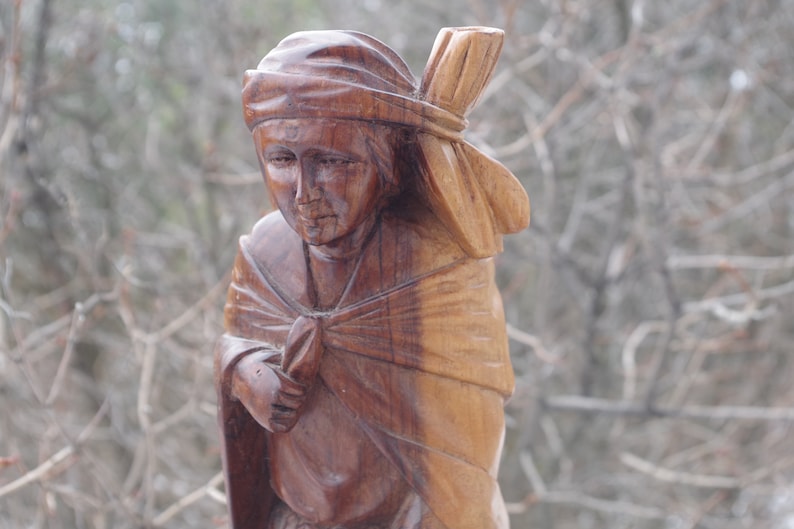Puede incluir: Una escultura de madera de una persona que lleva una cubierta para la cabeza y una capa. La escultura est&aacute; tallada en un estilo realista y tiene un color marr&oacute;n c&aacute;lido.