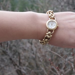 Könnte beinhalten: Eine goldfarbene Armbanduhr mit einem runden Zifferblatt und einem Gliederarmband.
