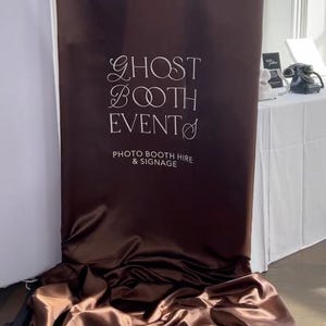 Puede incluir: Un telón de fondo de fotomatón de satén marrón con el texto blanco "GHOST BOOTH EVENT". También es visible el texto "PHOTO BOOTH HIRE & SIGNAGE". El telón de fondo está drapeado en el suelo.