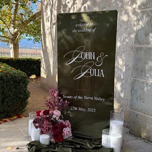 Puede incluir: Un letrero de bienvenida de boda verde oliva con la inscripción blanca "welcome to the wedding of John & Julia". Decorado con flores, velas y tela drapeada. La fecha es el 25 de mayo de 2025. El lugar es Stones of the Yarra Valley.