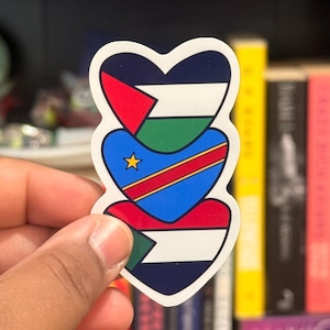 May include: A sticker with three heart-shaped designs, each with a flag pattern. The top heart has red, white, green, and black, the middle has blue, yellow, and red, and the bottom has green, white, and black. The sticker is held in a hand.