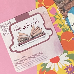 May include: A pink and yellow floral patterned bookmark with a pink and white book illustration and the Arabic text "رَّبِّ زِدْنِي عِلْمًا."  The text "Oh my Lord, advance me in knowledge." is written below the illustration. The bookmark is labeled "GEEKAABI'S WORLD MAGNETIC BOOKMARK".