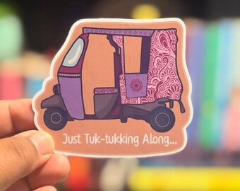 May include: A sticker featuring a purple and brown tuk-tuk with a decorative floral design. The sticker has the text "Just Tuk-tukking Along..." in a playful font. The background is blurred, suggesting the sticker is being held.