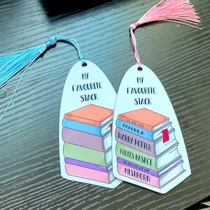Puede incluir: Dos marcadores de libros coloridos con una pila de libros y el texto "My Favorite Stack". Los marcadores son de forma ovalada y tienen un borla en la parte superior.