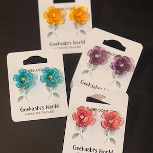 Puede incluir: Cuatro pares de pendientes con forma de flor en amarillo, turquesa, morado y rojo. Cada par se muestra en una tarjeta blanca con el texto "Geekaabi's World HANDMADE TREASURES" y "EVERBLOOMS".