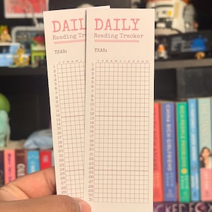 Może przedstawiać: Dwie białe karty śledzenia czytania z różowym napisem "DAILY Reading Tracker". Karty mają siatkę do śledzenia codziennego czytania, z miejscem na rok i miesiące. Karty są trzymane w dłoni.