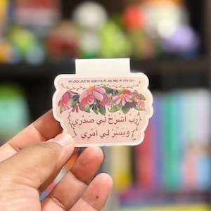 May include: A light pink sticker with floral accents and Arabic text. The sticker has the English text "MY LORD! UPLIFT MY HEART FOR ME AND MAKE MY TASK EASY" at the top. The sticker is held in a hand.