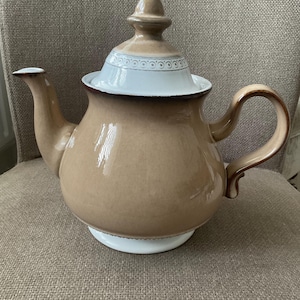 以下が含まれることがあります： 白とベージュの陶器のティーポット。蓋には装飾的な縁と小さなつまみがあります。ハンドルと注ぎ口もベージュです。ティーポットはニュートラルカラーの表面に置かれています。