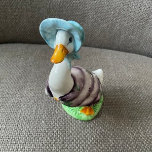 以下が含まれることがあります： 青い帽子と紫色のドレスを着たアヒルのセラミック製フィギュア。アヒルは緑色の土台の上に立っています。