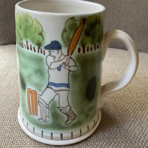 May include: Cream-colored ceramic mug with a hand-painted cricket scene. The design features a batter in a blue cap and white uniform, swinging a bat. Green trees and a cricket pitch are in the background. The mug has a curved handle and a decorative base.
