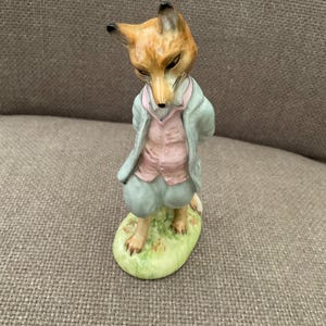 Puede incluir: Una figura de porcelana de un zorro de pie sobre una base verde. El zorro está antropomorfizado, viste una chaqueta azul claro, un chaleco rosa y pantalones cortos azul claro. Tiene pelaje marrón, orejas puntiagudas y mira hacia abajo.