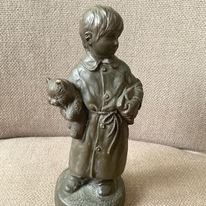 May include: A bronze-toned statue depicting a child clutching a cat and a book. The child is dressed in a coat with a rope tie, standing on a circular base. The statue has a detailed finish and is set against a neutral backdrop.