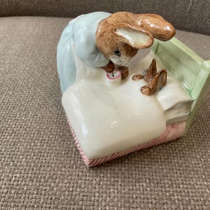 Peut inclure: Une figurine en céramique représentant un lapin brun vêtu d'une veste bleu clair, penché sur un lit pour border un plus petit lapin brun. Le lit présente une tête de lit vert pâle, une couverture blanche et une base à rayures roses et blanches.