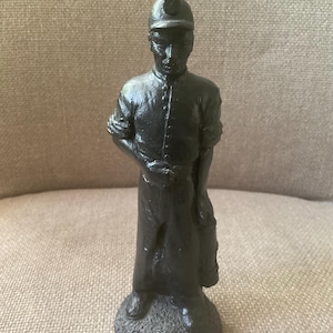 May include: A black figurine depicting a coal miner, standing on a textured base. The miner is wearing a cap, button-down shirt, and apron, and is holding a lantern. The figurine is a detailed representation of a historical occupation.