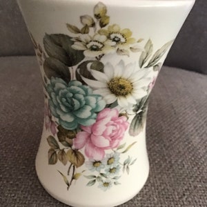 Könnte beinhalten: Eine weiße Keramikvase mit einem Blumenmuster. Die Vase hat eine breite Öffnung und einen schmalen Boden. Die Blumen sind in den Farben Rosa, Blau, Grün und Gelb.