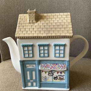 Op de afbeelding: Een keramische theepot in de vorm van een winkel met twee verdiepingen, een beige pannendak en een schoorsteen. De gevel is wit en blauw met drie ramen, een blauwe deur met de tekst Welcome en een etalage met thee, taart en vlaggetjes.