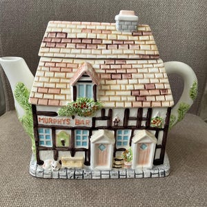Op de afbeelding: Een keramische theepot in de vorm van een pub, met een dak met baksteenmotief, een schoorsteen en de tekst "Murphys Bar". De theepot heeft een witte tuit en handvat, met groene bladaccenten. De gevel bevat deuren, ramen en decoratieve elementen.
