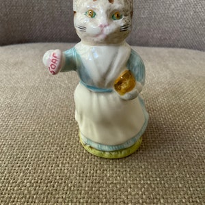 Puede incluir: Una figurita de cerámica de un gato con un vestido azul y sosteniendo un pan en una mano y un objeto en forma de corazón que dice "SOAP" en la otra.