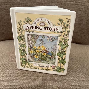 May include: A white ceramic book-shaped vase with a yellow and green floral design. The vase features the text "Brambly Hedge Spring Story" and "Jill Barklem". The illustration depicts a spring scene with flowers and a small creature.