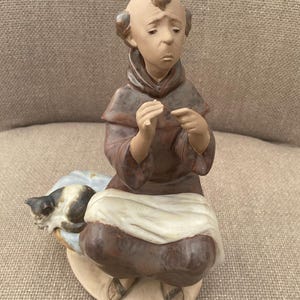 May include: A ceramic figurine of a monk in brown robes, seated with a sad expression. A small cat rests on a pillow beside him. The figurine has a detailed, hand-painted appearance.