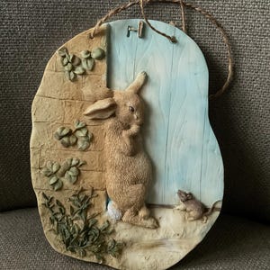 Puede incluir: Una decoración de pared de cerámica con un conejo marrón asomándose por una puerta azul, con un pequeño ratón en el suelo. La decoración de pared está unida a una cuerda de yute marrón.