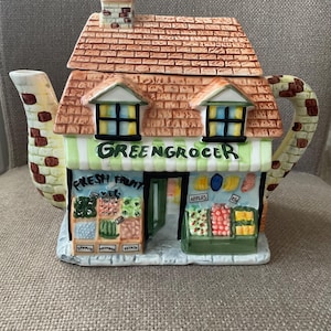 Op de afbeelding: Een keramische theepot in de vorm van een groentewinkel. Het dak is baksteenpatroon, met een kleine schoorsteen. De winkelgevel is voorzien van de tekst "GREENGROCER" en toont displays van vers fruit en groenten. De tuit en het handvat hebben een gestructureerd ontwerp.