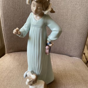 Könnte beinhalten: Porzellanfigur eines jungen Mädchens in einem hellblauen Kleid mit Zöpfen. Sie hält ein kleines Spielzeug und wird von einer kleinen Hundefigur begleitet. Die Figur ist detailliert und hat eine weiche, pastellfarbene Farbpalette.
