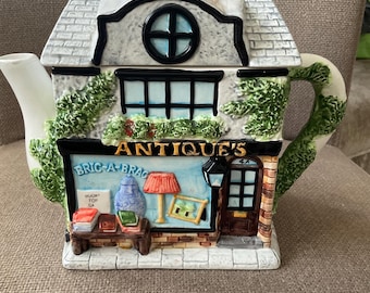 May include: A ceramic teapot in the shape of a two-storey building with a brick chimney. The building is labelled "ANTIQUE'S" and "BRIC-A-BRAC" with a shop front displaying miniature items. The roof is white with grey shingles, and the handle and spout are white.