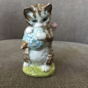 Puede incluir: Una figurita de porcelana de un gatito tabby marrón y blanco que sostiene una bola de lana a cuadros azules y blancos. El gatito tiene ojos verdes y la nariz rosa. La figurita está de pie sobre una base verde.
